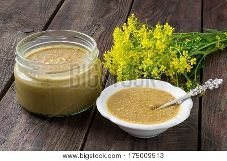Homemade mustard with grains in jar and gravy boat mustard flowers on old wooden table. Spicy seasoning for various dishes. Selective focus