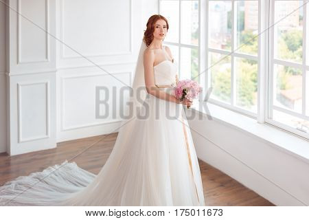 Bride in white wedding dress and long veil with bouquet of flowers indoors. Young gorgeous fashion model posing in white interior studio's room near big window in full lenght