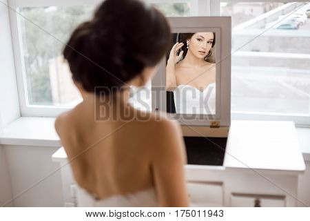 Portrait of young beautiful bride preparing to her wedding day. Gorgeous woman looking at mirror at her make-up and hairstyle