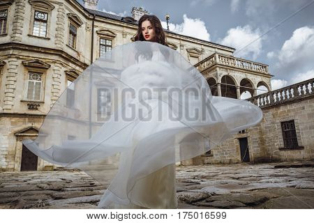 Young sexy girl with long hair and pretty face in white wedding bride dress posing near castle or palace building