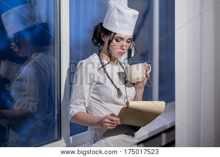 Brunette female nurse in uniform drinking tea and reading notes