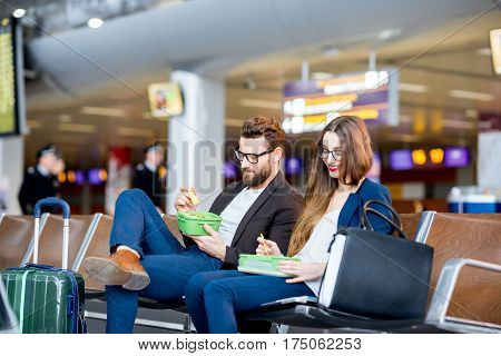 Elegant business couple eating with lunch boxes sitting at the waiting hall in the airport. Having a snack during business trip