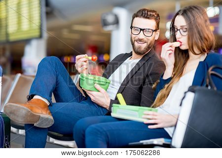 Elegant business couple eating with lunch boxes sitting at the waiting hall in the airport. Having a snack during business trip