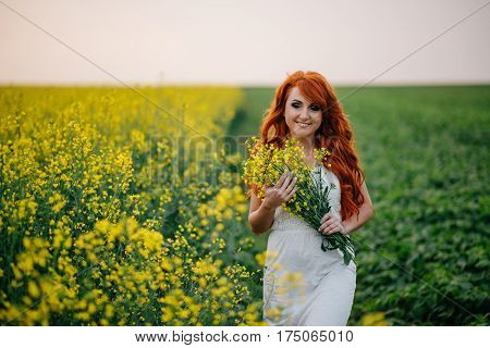 Young beautiful red-haired woman in a blooming rapeseed field at sunset