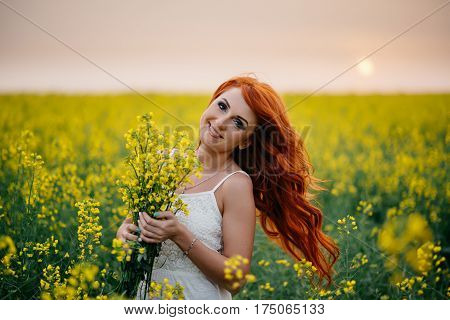 Young beautiful red-haired woman in a blooming rapeseed field at sunset