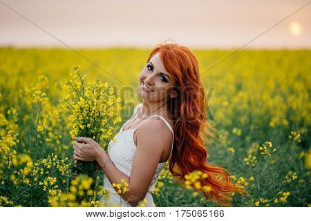 Young beautiful red-haired woman in a blooming rapeseed field at sunset