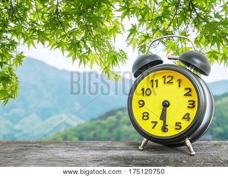 Closeup black and yellow alarm clock for decorate show half past six or 6:30 a.m. on blurred leaves and mountain view background