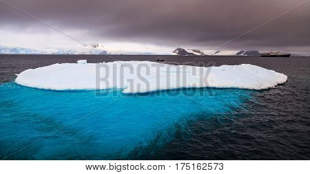 Massive iceberg with blue uderwater part and cruise ship in background Peterman Island Antarctica