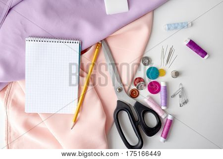 needlework and tailoring concept - scissors, sewing buttons, spools of thread, cloth and notepad with pencil