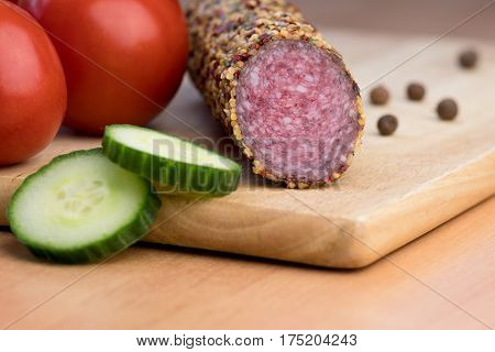 Delicious smoked sausage whole on wooden board with slices of cucumbers and tomatoes. View from above.