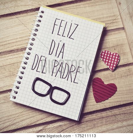 the text feliz dia del padre, happy fathers day in spanish written in the page of a notebook, a pair of black eyeglasses and some red hearts on a rustic wooden background
