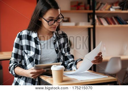 Picture of pretty young caucasian woman wearing glasses sitting indoors while using tablet computer and holding documents. Coworking concept.