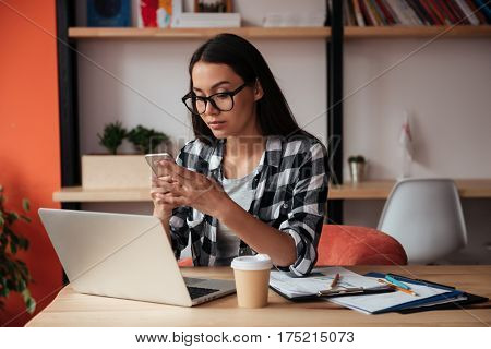Image of beautiful young caucasian lady wearing glasses sitting indoors while using mobile phone and laptop computer. Coworking concept.