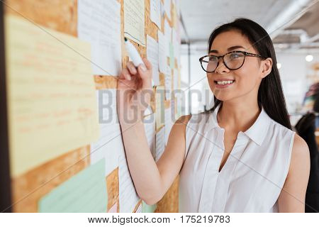 Happy pretty asian young woman in glasses writing advertisement on bulletin board