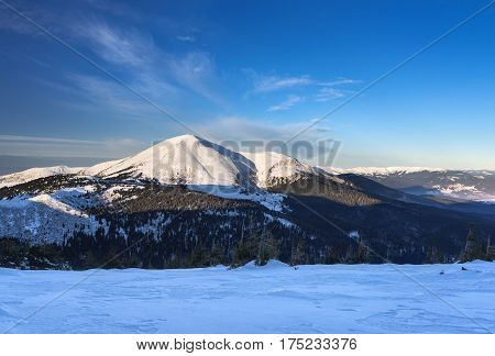 View at high mountain peak covered with snow. Forest at the bottom of the mountain and blue sky over the mountain