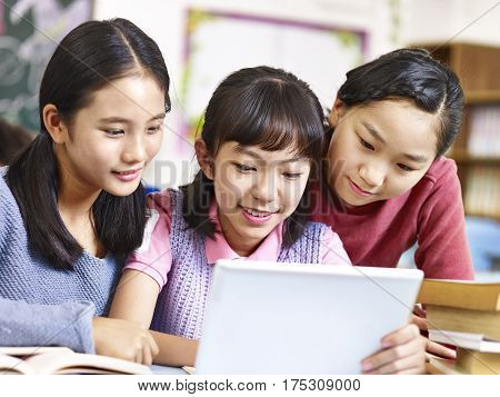 three asian elementary school girls friends looking at a tablet together during break in classroom.