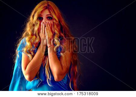 Frightened woman screaming with fear indoor at halloween night. Horror. Girl with tousled long blond hair and opened her eyes wide bawl covered her face on black background.