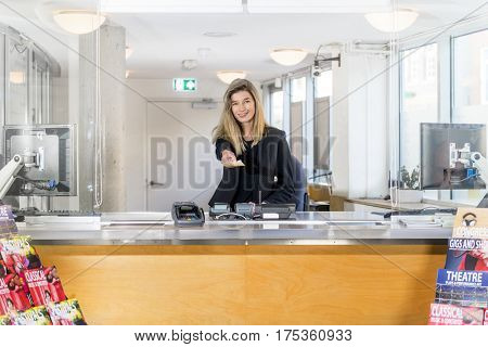 Saleswoman, reaching out her hands with tickets for an event at a theater, sitting behind the counter of a ticket booth, flyers of various performances in front of the sales office