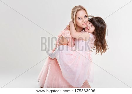 blonde mom and sweet little daughter in pink dresses princesses hugging and laughing on gray background. family