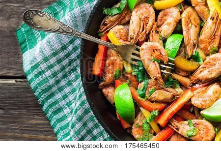 Fried shrimps with bell peppers cilantro and lime in iron skillet on rustic wooden background. Top view.