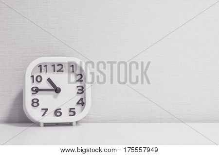 Closeup white clock for decorate show a quarter to eleven o'clock or 10:45 a.m. on white wood desk and cream wallpaper textured background in black and white tone with copy space