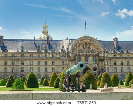 Part of the northern facade of the Hotel Des Invalides with archway of a main entrance with low-relief of King of France Louis XIV bronze cannon and garden in the foreground in Paris