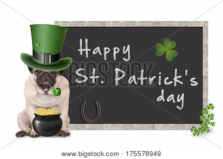 cute pug puppy dog with leprechaun hat for st. patrick's day smoking pipe sitting next to blank blackboard sign with horseshoe and shamrock on white background