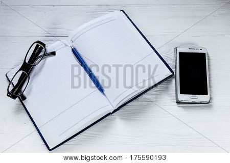 Blank notebook with pen. eyeglasses and mobile phone on wooden table, business concept