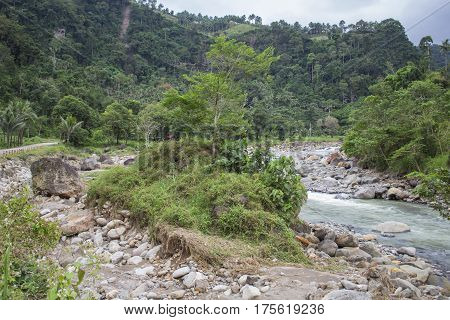 Tropical landscape with small river in stone riverbed and forest hills. Rocky riverbed with small island. Tropic greenery in rainy season. Jungle bush and trees in river valley between mountains.