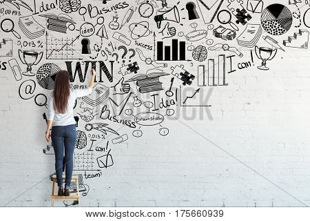 Back view of young woman standing on stool and drawing creative business sketch on brick wall. Leadership concept