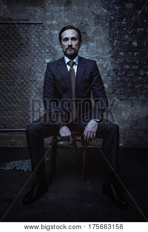 Super villain. Good looking brunette sharp gentleman wearing a classic suit and sitting on a chair in a dark room while holding a gun