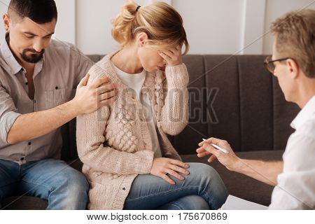 Being at loss. Desperate beautiful blonde woman sitting near her husband and holding her forehead while thinking about her problems