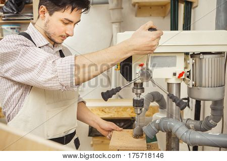 Brunette man bored wooden board on drilling machine. Woodworking drill installed on machine with engine. Mounted hoods to place of drilling.