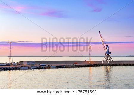 Multi-ton industrial port crane on rails lifts heavy loads in marine passenger-cargo harbour of Tallinn. Evening panorama of sea bay at sunset. Concrete seawall with lighting masts & crane. Estonia
