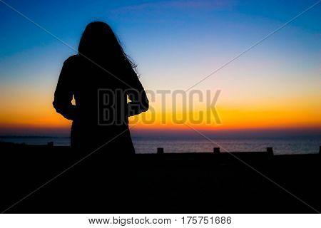 The silhouette of a young woman looks out to sea while the sun goes down in a colourful haze