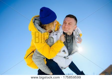 Young man holding his girlfriend on his shoulders. Girl embraces guy for neck. Young people look at each other with love and tenderness. Against the background of the blue sky.