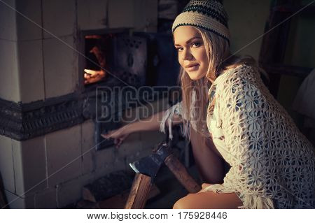 Young blonde woman spliting logs and burning wood in stove at home