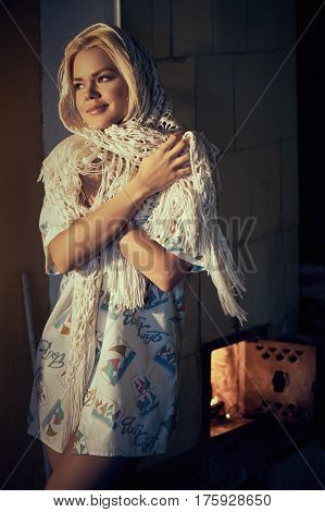 Young blonde beautiful woman in kerchief basking near heating stove at own home