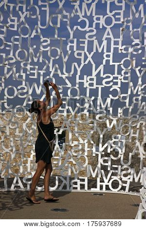 Young beautiful woman during shooting. Composition with white letters on a blue sky. Detail of sculpture Nomad 2010. Painted stainless steel by Jaume Plensa (Barcelona Spain). Antibes Cote d'Azur France. June 30 2016