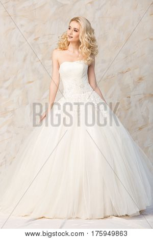 Portrait of the gorgeous smiling blonde bride with beautiful curly hair in a white wedding dress to the full-length on a light background. beautiful young model with bridal hairstyle and makeup in a white magnificent dress.