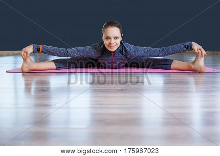 autiful young woman wearing blue sportswear set working out against blue wall, doing yoga or pilates exercise. Woman sitting splits. On the pink yoga mat. In a gym.