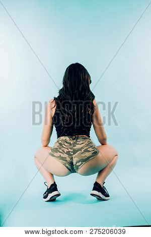 Sexy Brunette Woman Dancing Twerk On Blue Background, From Behind. A Young Girl Wears Camo Shorts An