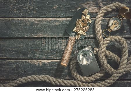Compas, Gold Nuggets On The Spade, Rope And Vintage Flashlight On The Aged Wooden Table Background W