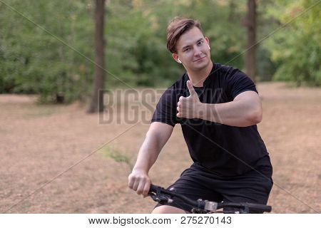 Young Handsome Caucasian Man Thumbs Up Bicycling At The Park. White Earphones, Dark Casual Wear. Out