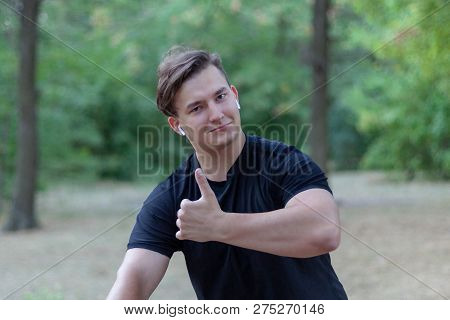 Handsome Young Caucasian Man Thumbs Up The Park. White Earphones, Dark Casual Wear. Outdoors, Abando
