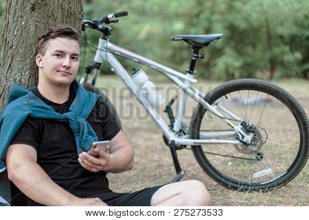 Attractive Young Caucasian Man Slightly Smiling, Holds Mobile Phone, Sitting On The Ground Close To 