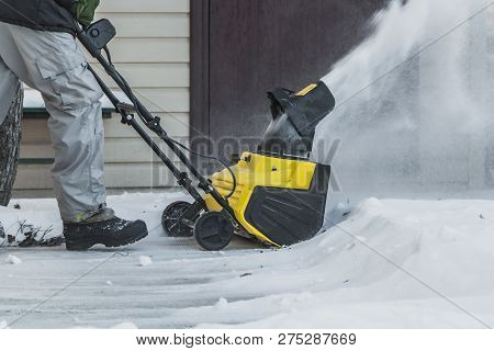A Man In A Black Jacket And A Gray Pants Is Brushing White Snow With The Yellow Electric Snow Throwe