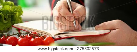 Male Cook Hand Hold Siver Pen And Wright Open Book Of Recipes On Background Of Vegetables. Chef Writ