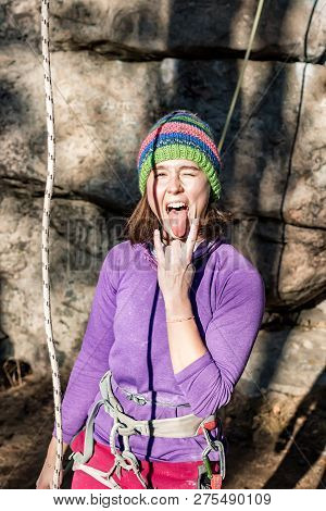 Portrait Of Girl Rock Climber Shows Tongue Happy And Cheerful, Standing At Rock Inharness With Rope 