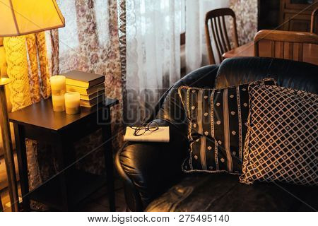 Photo of books, candles and glasses on a coffee tray by the sofa in log cabin. Cozy place for reading and relaxing some cold winter or autumn evening.